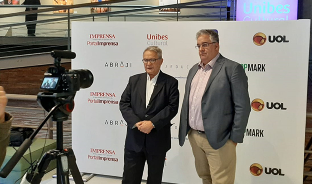 Dois homens executivos posando para foto, no fundo um banner de fundo branco com logo dos patrocinadores