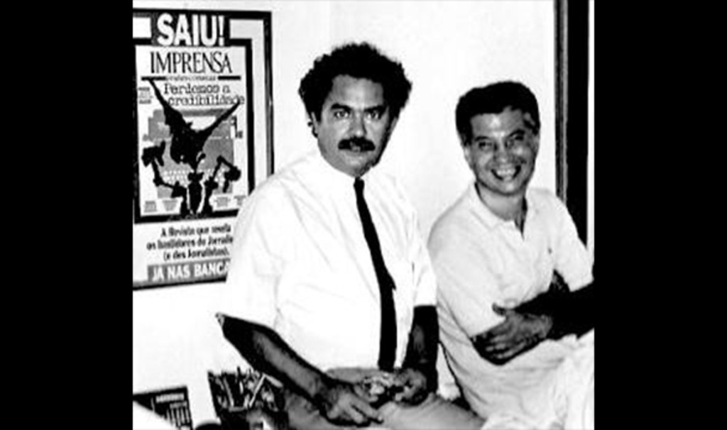 Foto preto e branca antiga de dois homens vestindo camisa clara num escritório, ao fundo um quadro divulgando a revista IMPRENSA