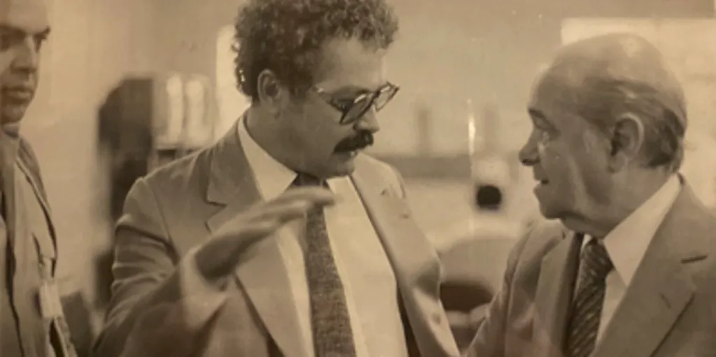 Foto em preto e branco mostra homens de terno conversando. O homem ao centro gesticula com a mão enquanto fala com o homem à direita; ambos parecem envolvidos na conversa.